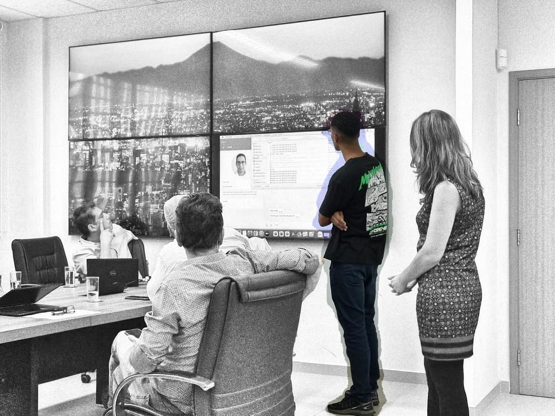 Uma reunião de trabalho em uma sala de conferências, onde cinco pessoas estão discutindo uma apresentação exibida em uma grande tela na parede. A tela mostra uma cidade iluminada ao entardecer e uma interface de software com a foto e informações de uma pessoa. Um homem em pé, vestindo uma camiseta preta com estampas verdes e brancas, está olhando para a tela enquanto os outros participantes, sentados ao redor da mesa, observam atentamente. Uma mulher de vestido estampado também está em pé, próxima ao grupo. A sala tem paredes brancas, cadeiras executivas e notebooks sobre a mesa.